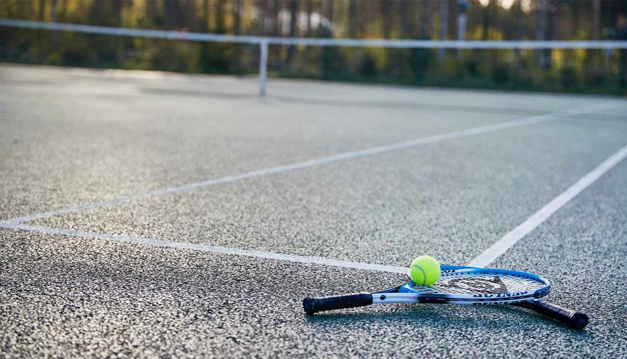 Two tennis rackets rest with a tennis ball balanced on one, lying on painted lines of an outdoor court; the net stretches behind, with sunlit trees blurred in the background.