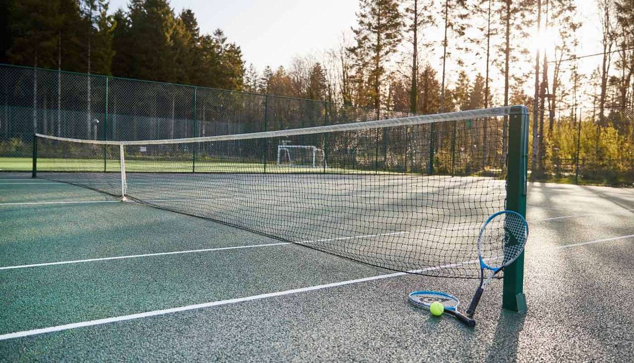 Two tennis rackets and a ball rest against a net post, on an empty outdoor court, surrounded by chain-link fencing and tall trees in warm, low sunlight.