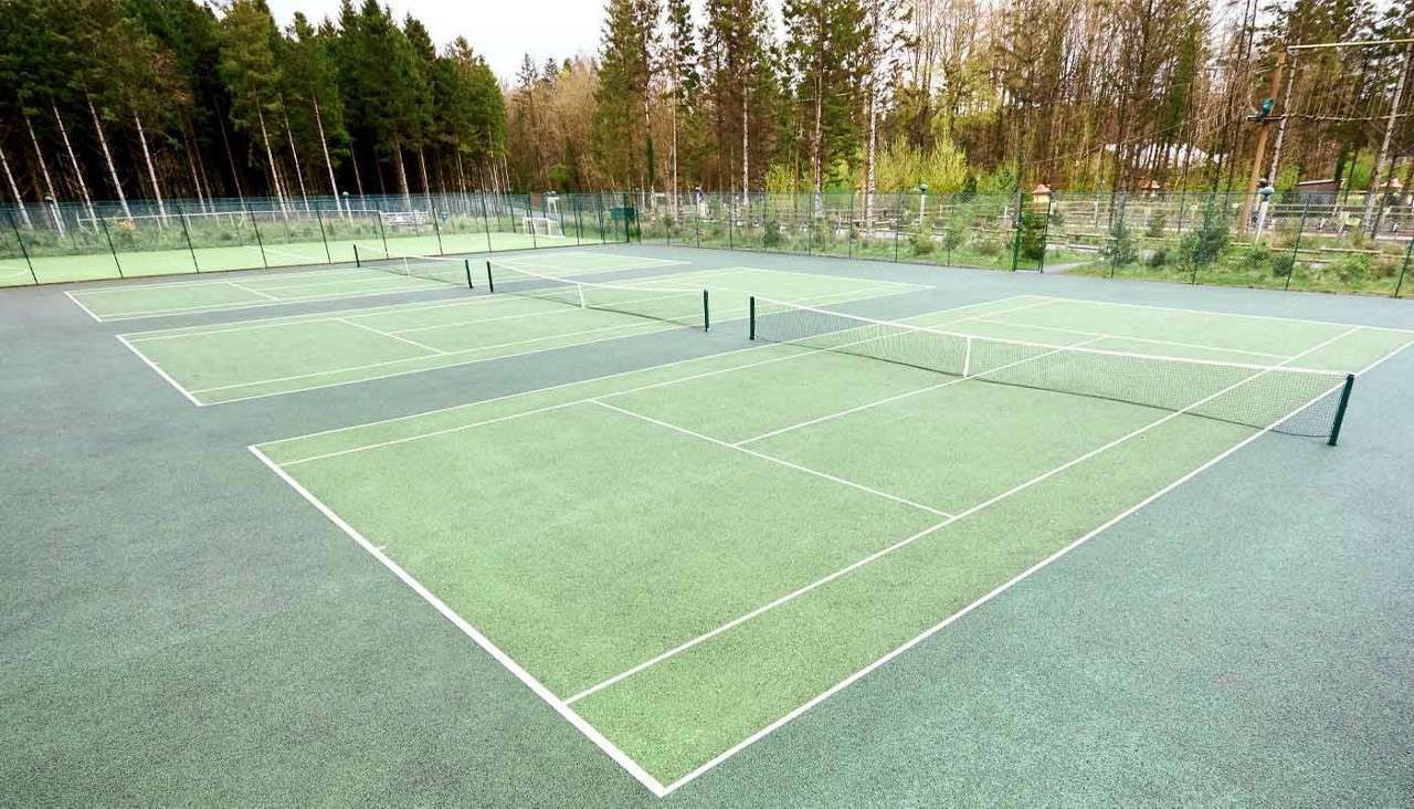 Multiple empty tennis courts sit side-by-side, nets stretched taut, within a fenced outdoor complex, surrounded by tall trees and greenery under overcast daylight.