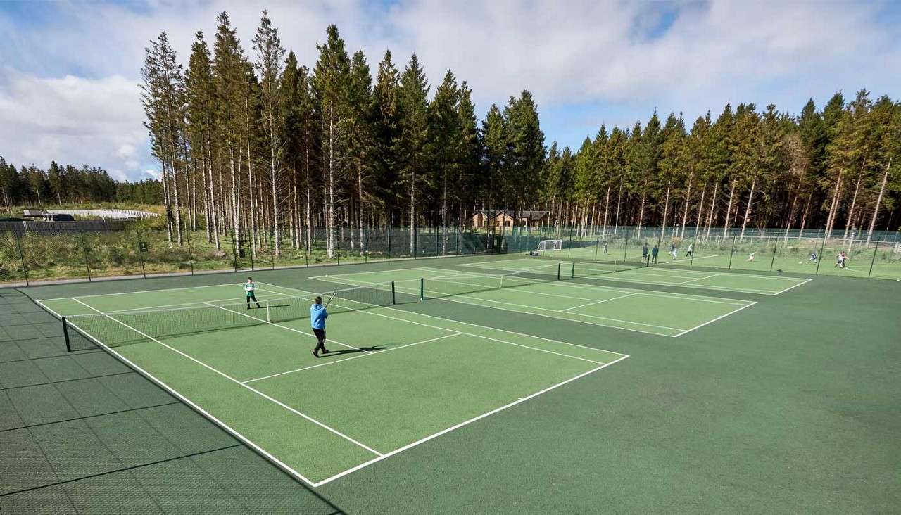Two tennis players rally on a green hard court, while others play on adjacent fenced courts, set outdoors amid tall pine trees under a partly cloudy sky.