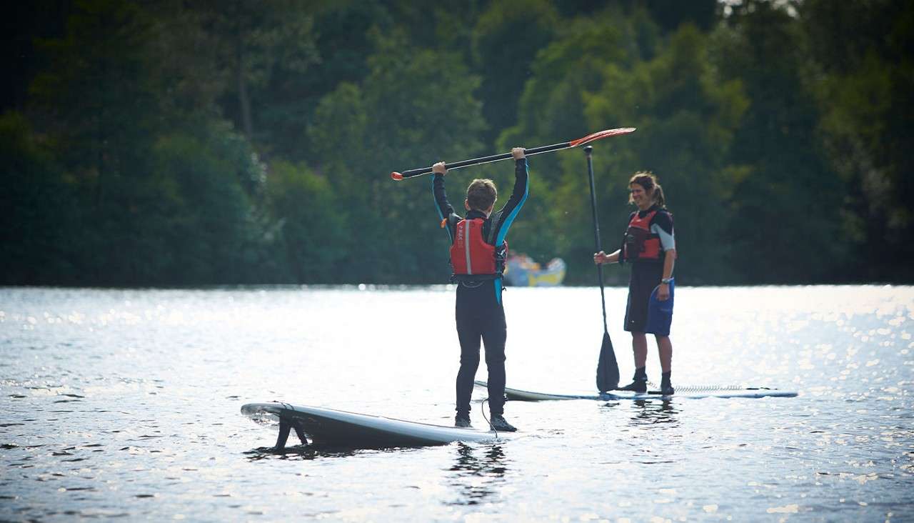 Two people on stand-up paddleboards; one raises a paddle overhead, the other stands holding a paddle, watching; calm lake water with ripples, life vests worn, forested shoreline in bright daylight.
