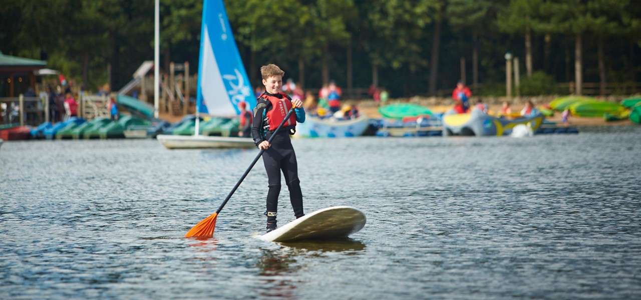 Child on a stand-up paddleboard paddles forward, wearing a wetsuit and red life jacket; behind, a lakeside activity center with kayaks, people, and a blue sail marked “Pico” near trees.