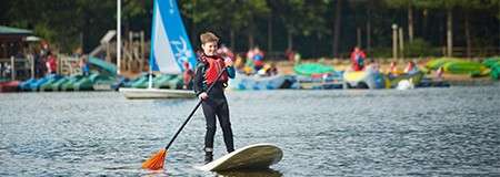 Person paddleboards upright, paddling with an orange-bladed oar on a calm lake; behind, sailboats, docks, and groups of people line a wooded shoreline in soft daylight.