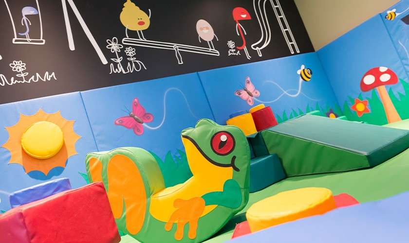 Frog-shaped soft play structure reclines on padded floor, surrounded by colorful foam blocks, ramps, and circles; in an indoor children's playroom with butterfly, bee, sun murals and chalk-style drawings above.