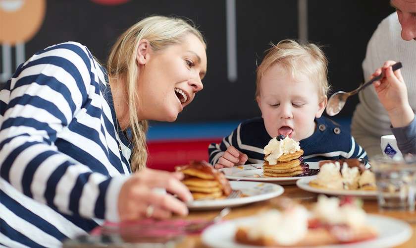 Toddler eats pancakes topped with whipped cream at table; a smiling woman leans close as another hand offers a spoon. Plates of pancakes and desserts sit on a restaurant table.