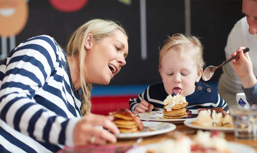 Toddler licks whipped cream atop a pancake stack while seated at a table, flanked by smiling adults, in a casual restaurant setting with other plates of pancakes.