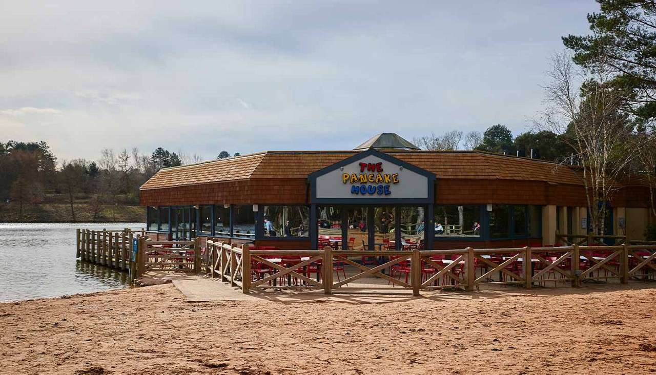 Lakeside restaurant displays sign “THE PANCAKE HOUSE,” seating sits empty, beside wooden deck over water, surrounded by sandy beach and trees under a cloudy sky.