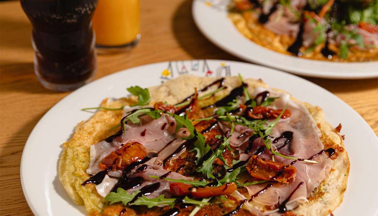 Savory crepe topped with prosciutto, arugula, sun-dried tomatoes, and balsamic drizzle rests on a white plate; behind it, cola and orange juice glasses flank another plate on a wooden table.