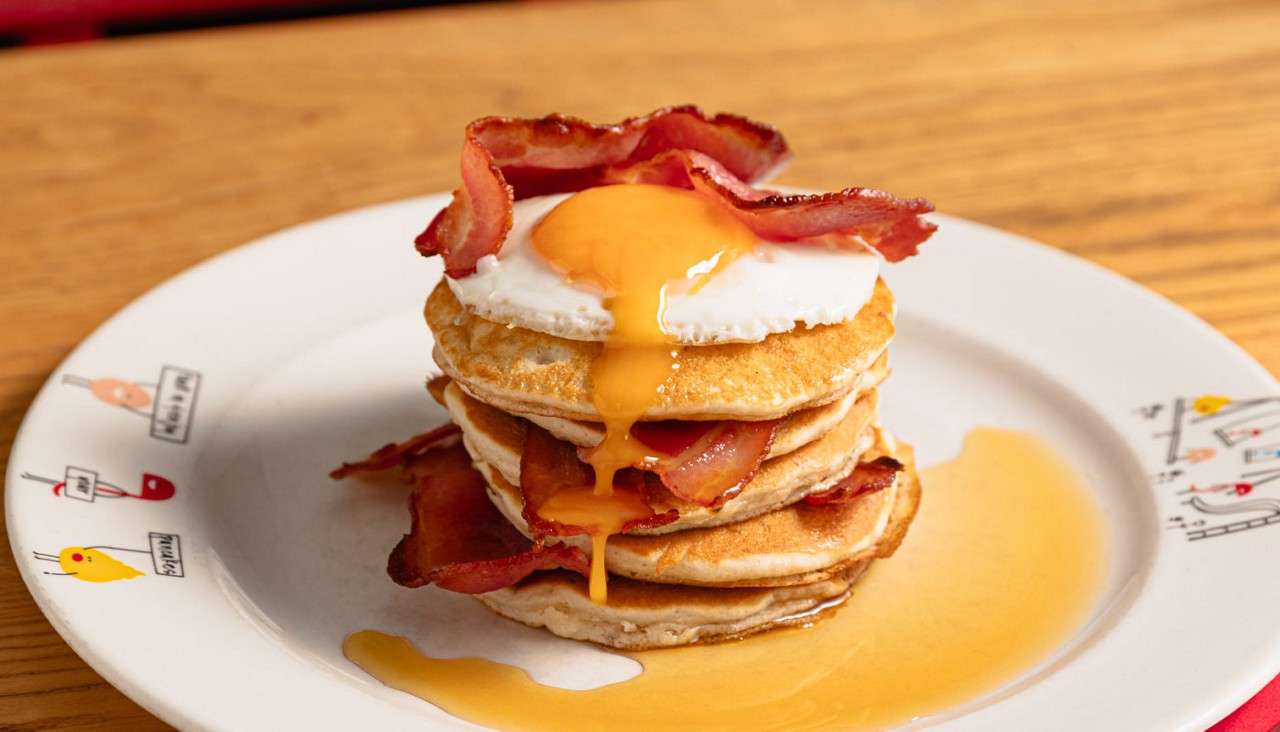 Stack of pancakes oozes runny fried-egg yolk and syrup, layered with crispy bacon, resting on a white patterned plate atop a wooden table in a casual dining setting.