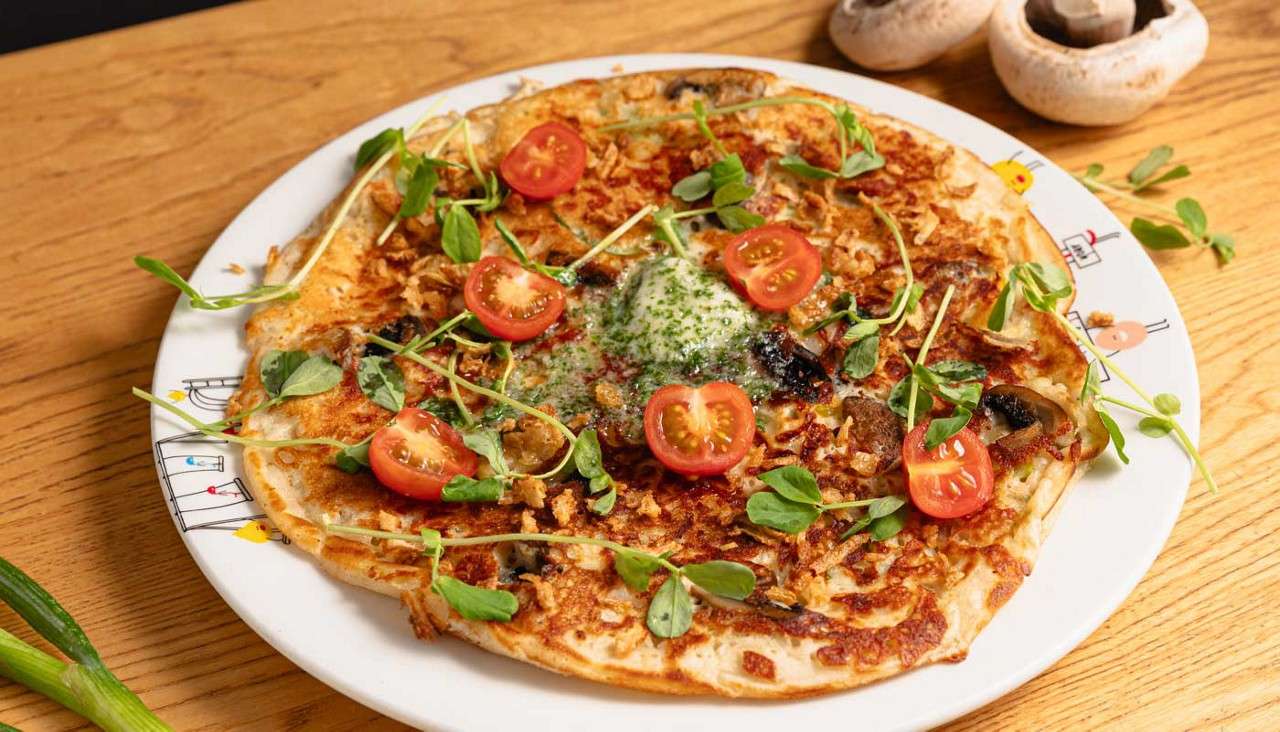 Savory pancake sits topped with halved cherry tomatoes, greens, and melted herb butter, sprinkled with crispy bits; presented on a white plate, on a wooden table beside mushrooms and scallions.