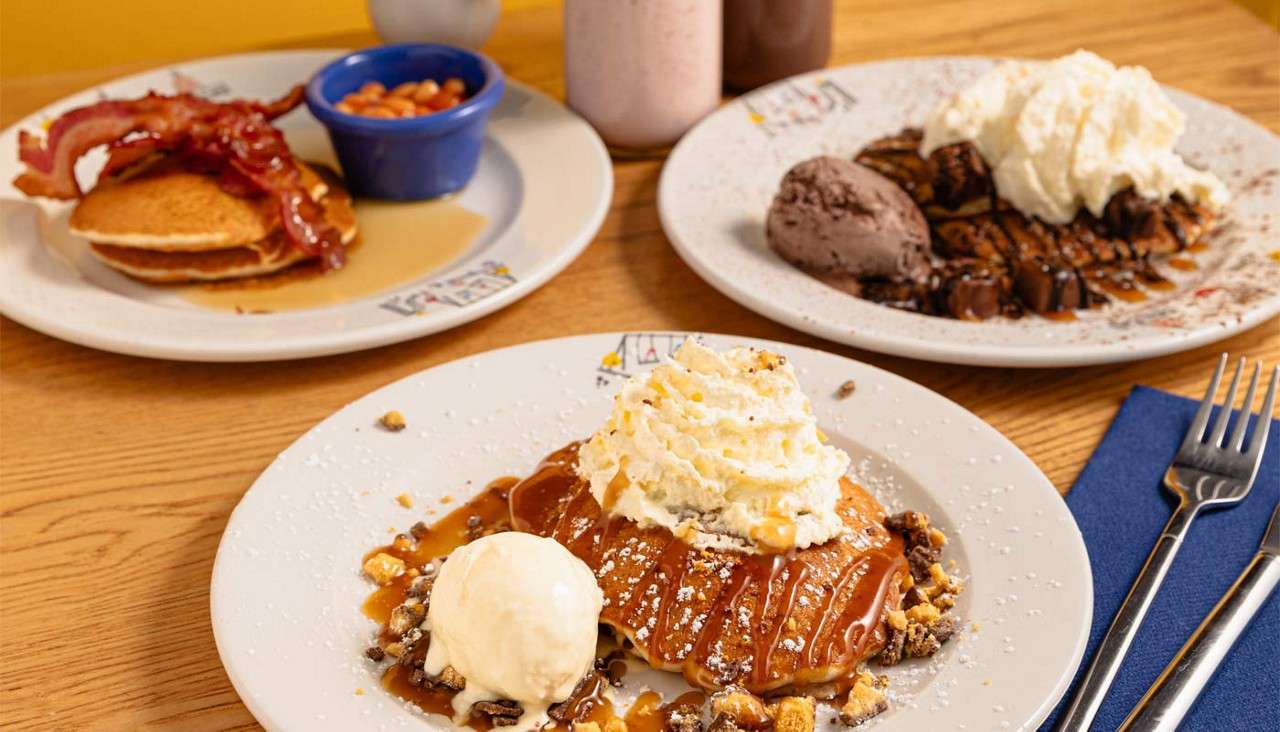 Pancakes topped with whipped cream and caramel, beside a scoop of ice cream. Other plates show bacon pancakes and chocolate waffles, with milkshakes, fork, and napkin on a wooden table.