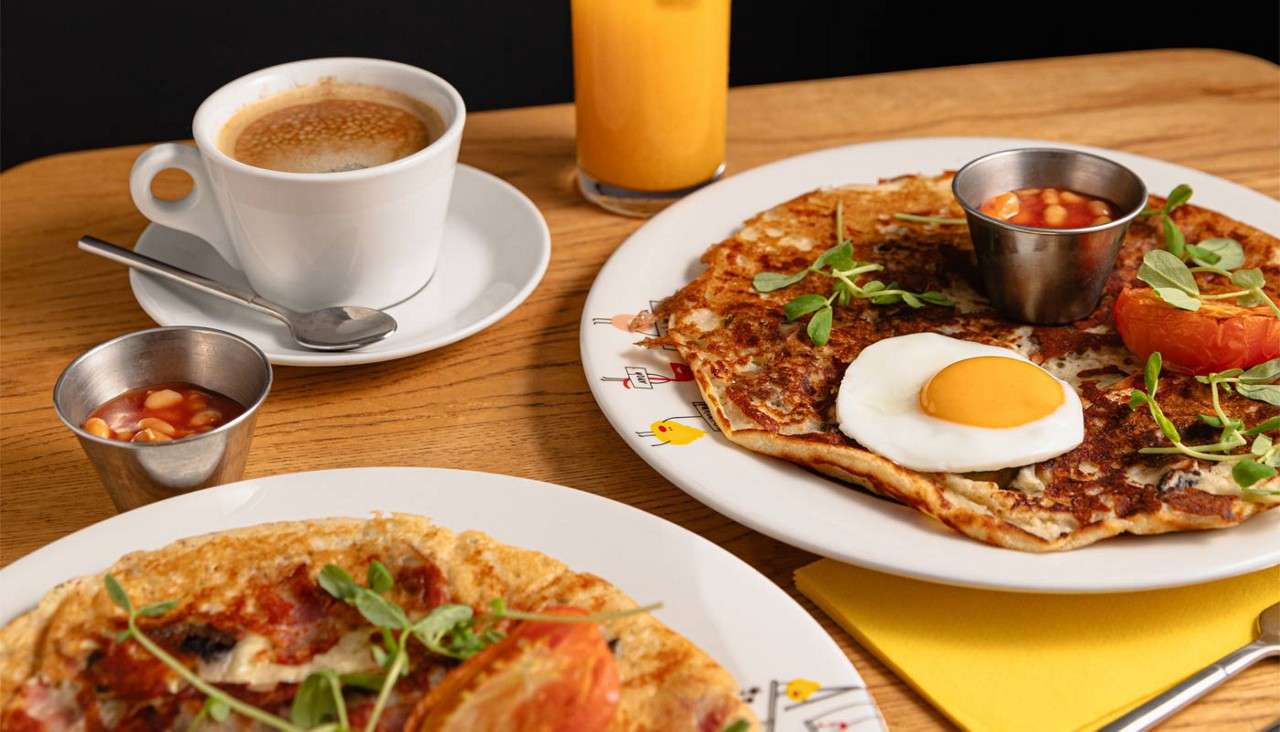 Breakfast plates sit on a wooden table; crepes topped with fried egg, tomato, greens, and baked beans. Nearby, a cup of coffee, orange juice, and metal ramekins accompany.