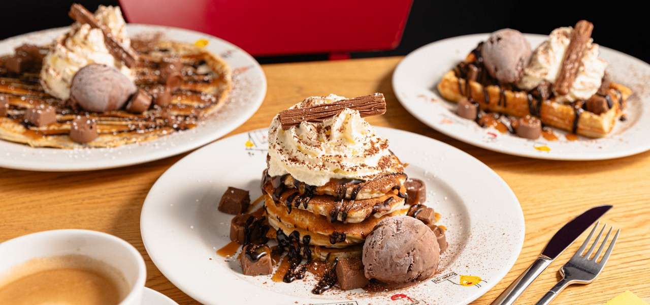 Pancake stack drizzled with chocolate and caramel, crowned with whipped cream and a chocolate flake, sits on a plate beside ice cream, waffles, coffee, and cutlery on a wooden table.