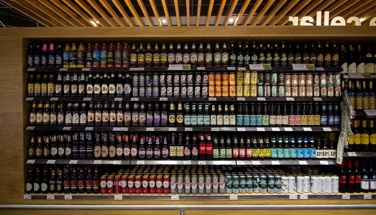 Bottles and cans stand neatly arranged on supermarket shelves, forming a colorful beer display under wooden ceiling slats. Visible text: £2, £5, IPA, 312.