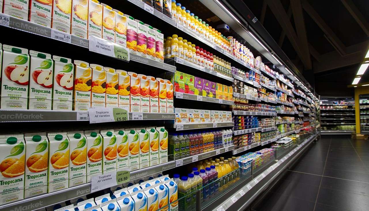 Juice cartons line refrigerated supermarket shelves. Text: “Tropicana,” “Original,” “Pressed Apple,” “Tropicana All Varieties,” “ANY 2 FOR £5.50,” “£3.45,” “ParcMarket,” “Naked.”