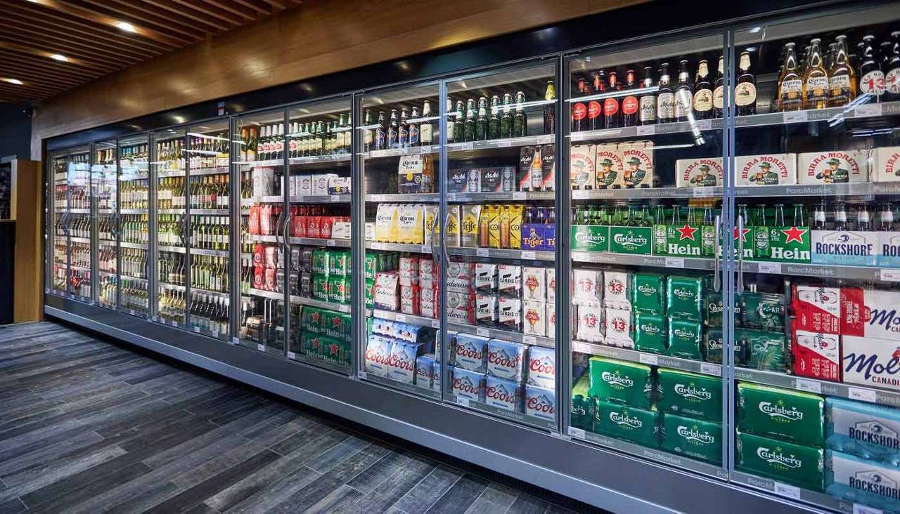 Glass-door refrigerated display holds assorted beer packs and bottles, arranged by brand, in a grocery aisle with wood ceiling and gray tile floor. Text: Carlsberg, Carlsberg Pilsner, Heineken, Rockshore, Birra Moretti, Corona, Asahi, Peroni, Tiger, Hop House 13, Coors, Coors Light, Molson Canadian.