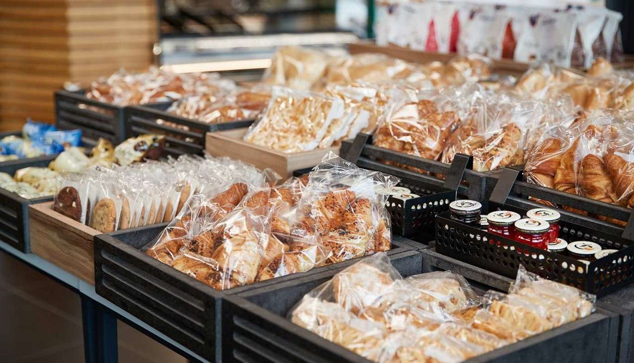 Packaged pastries sit in crates, arranged for sale on a bakery counter, with croissants, cookies, rolls, and small jam jars grouped neatly in a bright, self-serve display.