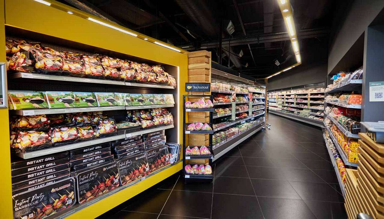Shelves display barbecue supplies and packaged produce, lining long aisles under bright lights in a modern supermarket. Text: BANANAS; INSTANT GRILL; Instant Party Barbecue; FLAMERS; BBQ; Allergens.