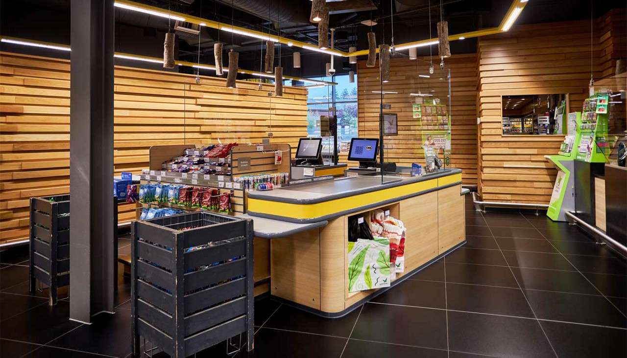 Checkout counter sits idle with two monitors and a conveyor belt; nearby candy racks and bins await shoppers in a modern wood-paneled grocery store with tile flooring and linear lights.
