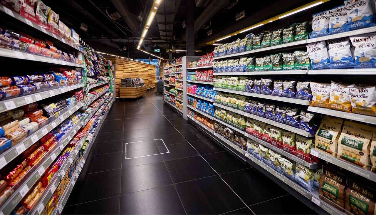 Snack shelves stand fully stocked, lining a supermarket aisle; chip bags and cookie packages face forward under bright lights, with a wooden island ahead. Legible text: “Keogh’s,” “Popcorn,” “CRINKLE.”