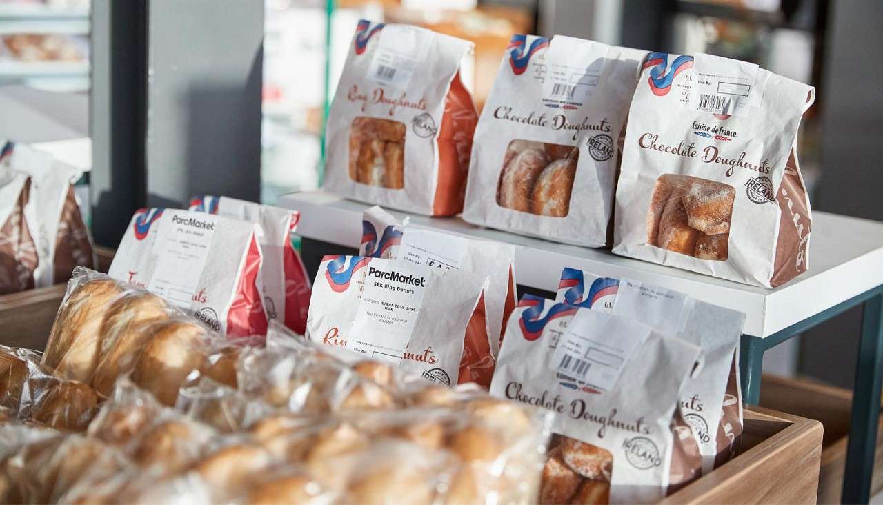 Bags of doughnuts sit on store shelves, their windows showing sugared rings, with wrapped bread rolls below in a wooden bin within a grocery bakery.

Text: Chocolate Doughnuts; Ring Doughnuts; ParcMarket; 5PK Ring Donuts; cuisine de france; IRELAND.