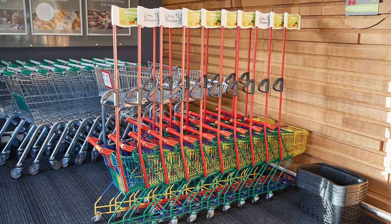 Children’s shopping carts stand nested, topped with red poles bearing small flags reading: Little Shopper. They sit near stacked full-size carts and plastic baskets in a grocery store entryway.