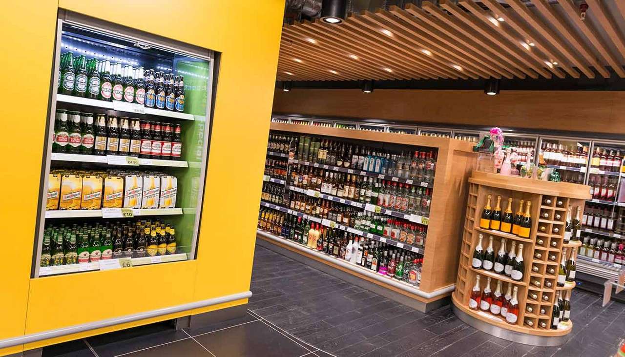 Refrigerated beer case displays bottles; adjacent shelves stock spirits in a modern supermarket. Text: Heineken; San Miguel; £10; £6.