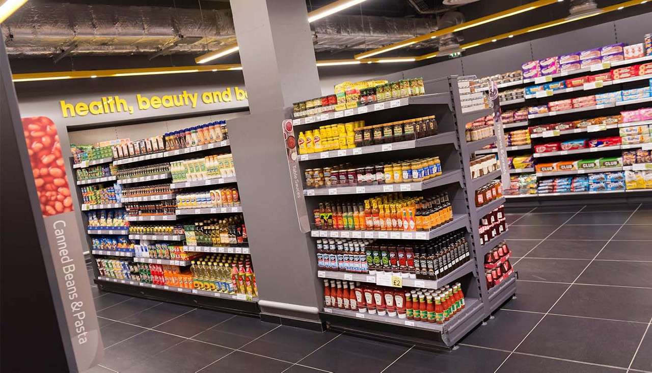 Shelving units display sauces, condiments, and snacks in a modern supermarket aisle with tiled floor and exposed ceiling ducts. Text: “health, beauty and b”, “Canned Beans & Pasta”, “£2”, “CLUB”, “HP”.