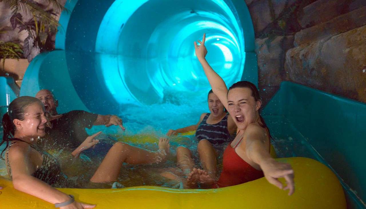 Group on a yellow inflatable raft rides out of a glowing blue tube waterslide, splashing and cheering, in an indoor waterpark with rock walls, plants, and rushing water.