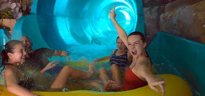 Group of four riders cheer and splash while seated on a yellow inflatable raft, emerging from a glowing blue tube waterslide inside an indoor water park with rock walls.