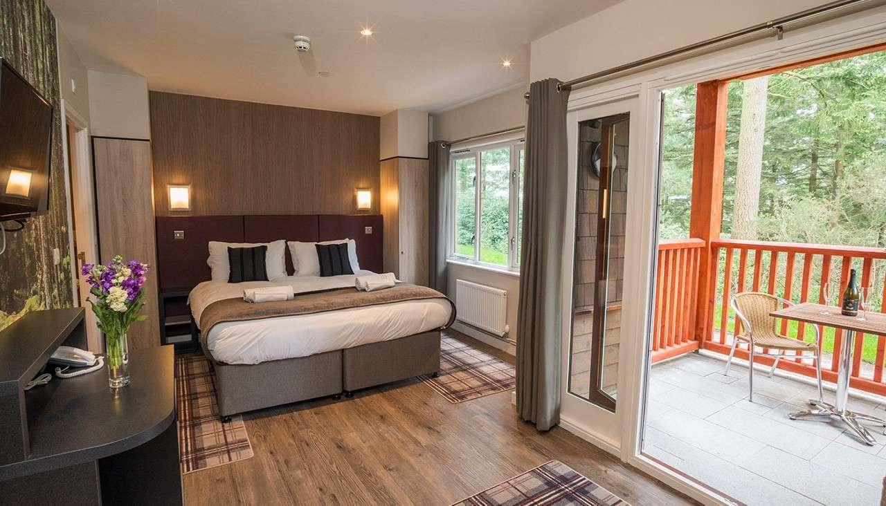 King bed rests neatly with pillows and towels, facing a wall-mounted TV. Sliding doors open to a balcony with table and chairs, overlooking pine trees in a serene lodge-style room.