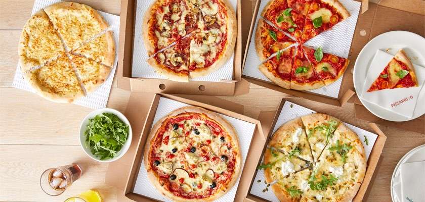 Assorted pizzas sit sliced in open boxes, some pieces removed. Surrounding them, a plate with a single slice, a small salad bowl, napkins, and drinks on a light wooden table.