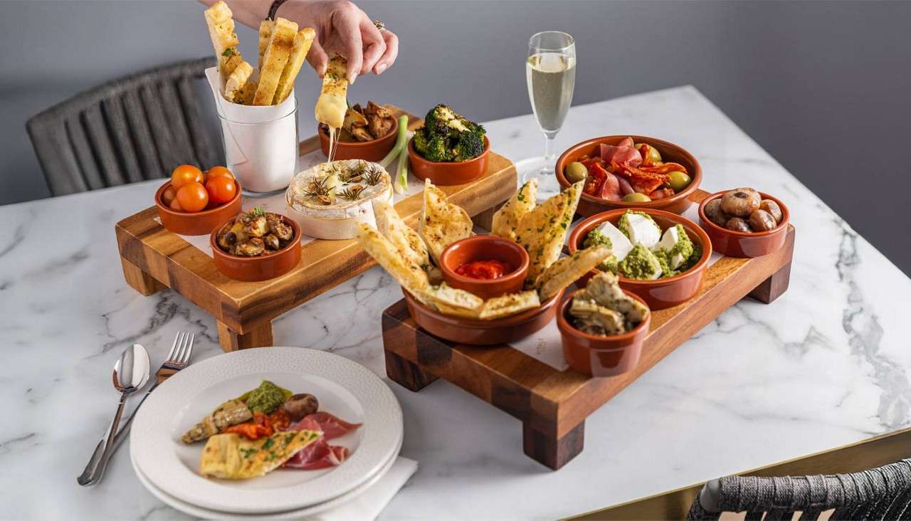 Tapas platters—breadsticks dipping into baked cheese—sit on wooden risers. Surrounding bowls hold olives, tomatoes, roasted vegetables, dips, and flatbread, beside a plated sampler and a glass of sparkling wine.