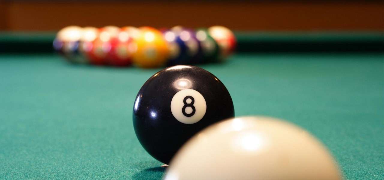 Eight ball marked "8" sits near a cue ball, both on a green billiards table, with a racked triangle of colorful balls blurred in the background.