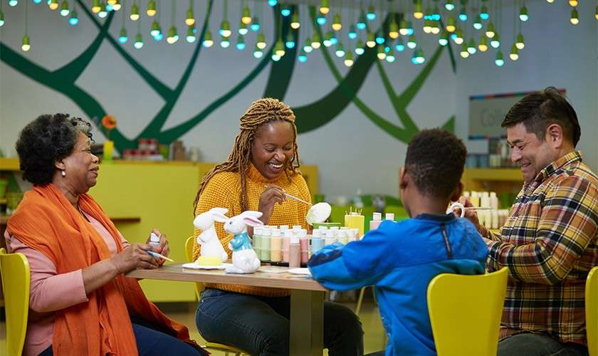 Group of adults and a child paint ceramic figures using brushes at a table in a craft studio, surrounded by glaze bottles, yellow chairs, and multicolored lights.