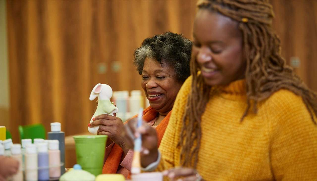 Two women paint ceramics; one smiles while holding a small mouse figurine, the other brushes glaze. Bottles of paint and cups surround them on a table in a craft studio.