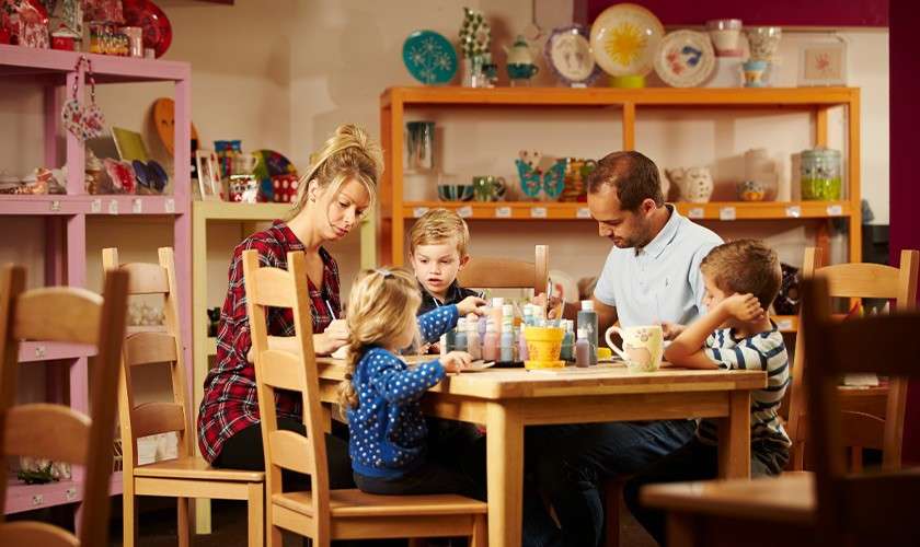 Family paints ceramics together at a wooden table, children and parents using brushes and paint bottles, surrounded by shelves of colorful pottery and craft supplies in a cozy studio.