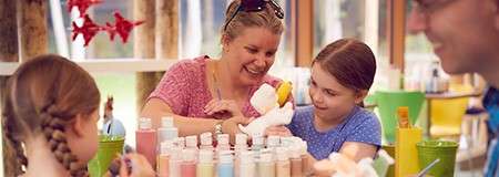 Children and an adult paint a ceramic figure with brushes, surrounded by rows of pastel paint bottles, in a bright, cheerful craft studio.