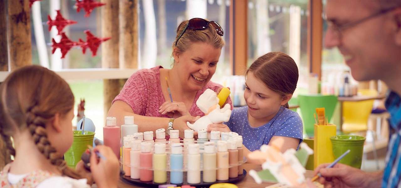 Family paints ceramic figurines at a table, using bottles of pastel paint on a rotating tray, in a bright craft studio with windows, green chairs, and hanging red paper stars.