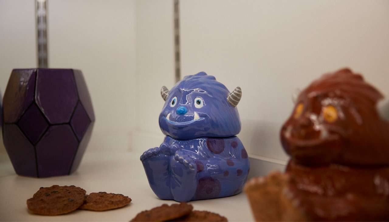Blue ceramic monster figurine sits grinning with feet forward on a white shelf, beside scattered cookies, a faceted purple container, and an out-of-focus brown monster figurine in the foreground.