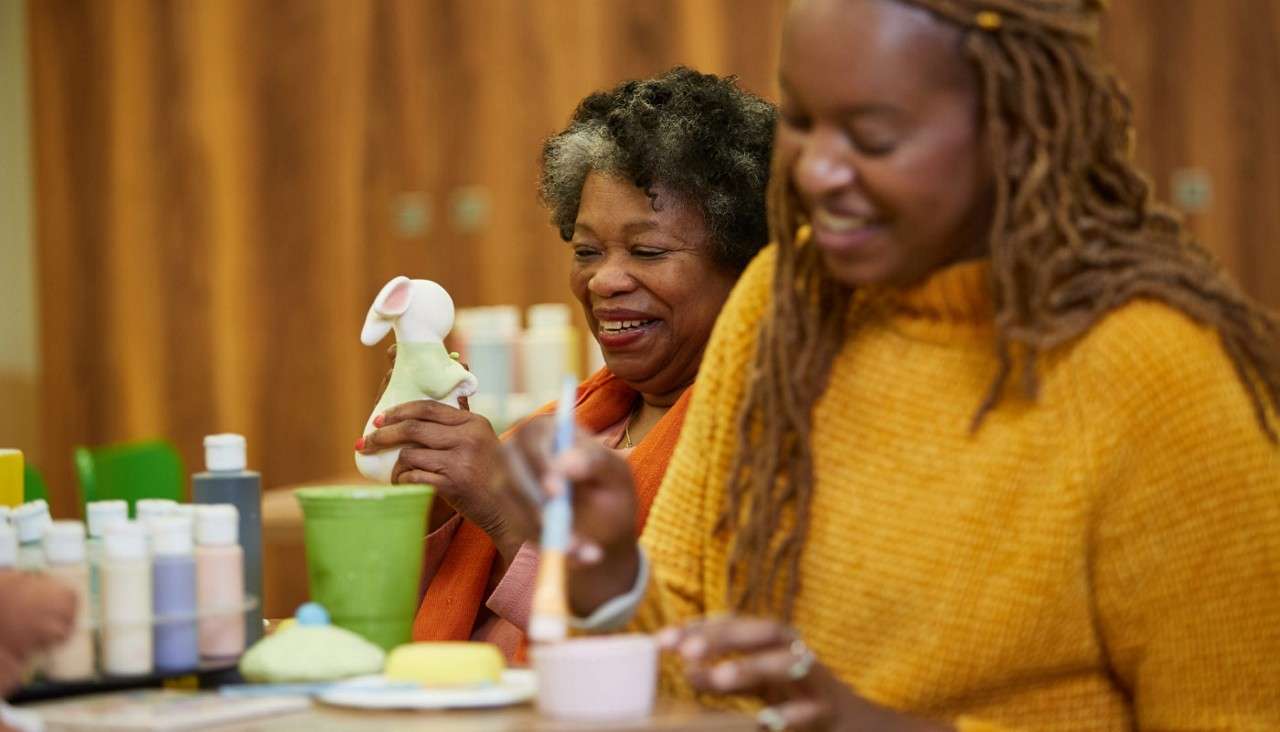 Two people paint ceramics; one holds a rabbit figurine while the other brushes a small cup, surrounded by paint bottles and tools on a table in a wood-paneled craft studio.
