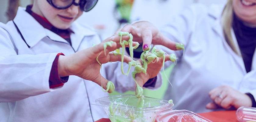 Child’s hands stretch sticky green slime, strands dangling between fingers, as an adult guides nearby; both wear white lab coats and safety goggles at a red-topped table in a classroom-like lab setting.