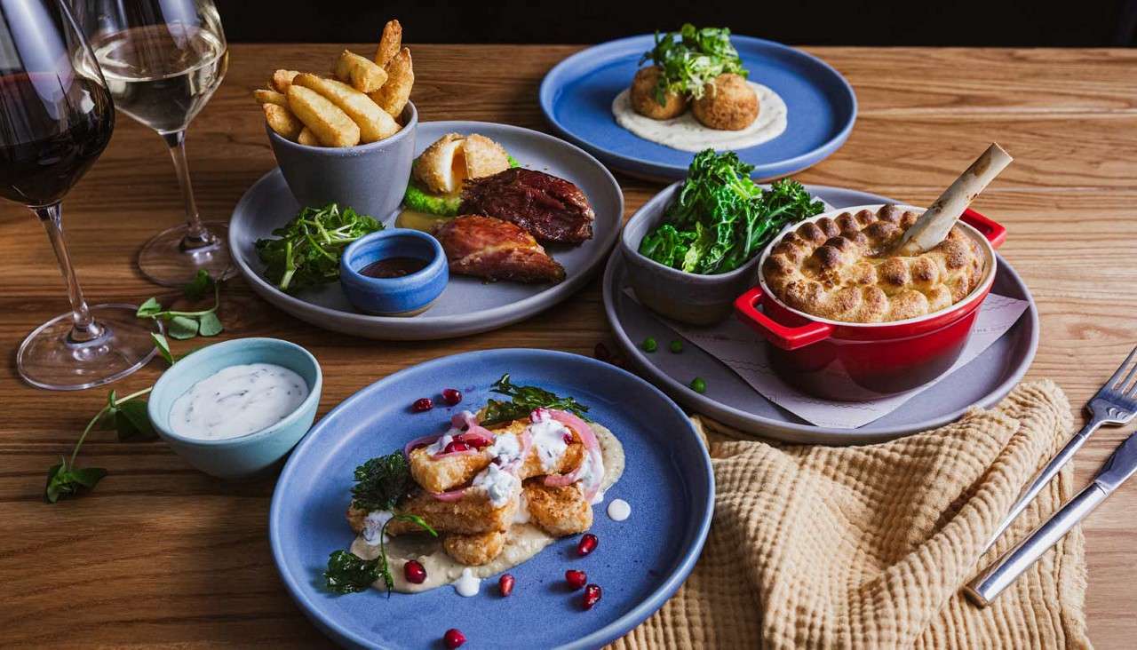 A multi-dish dinner is served, featuring fries, roasted meat, croquettes on puree, greens, nuggets, and a pie in a red pot, with wine, dips, and cutlery on a wooden table.