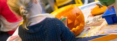 Pumpkin—being carved with a green-handled tool—sits on a newspaper-covered table; a child with braided hair works carefully indoors, with another small pumpkin and craft supplies nearby.