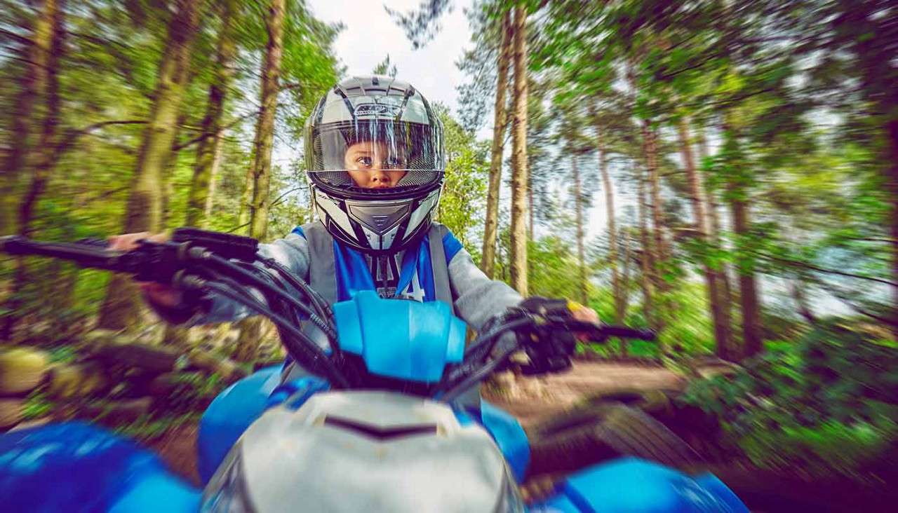 Child on a blue ATV rides forward, gripping handlebars, eyes visible through a full-face helmet. Surroundings blur into a green forest trail, suggesting speed. Visible text: 4.