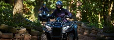 ATV rider steers along a narrow forest trail, navigating between stacked tires; another rider follows behind under dappled sunlight in dense woods.