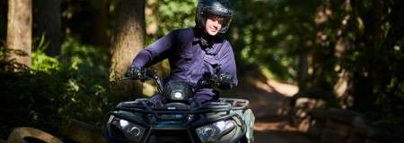 ATV rider steers along a shaded forest trail, wearing a helmet and dark clothing, navigating between trees with dappled sunlight on the path.