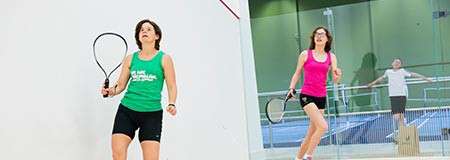 Two squash players swing racquets, chasing a shot inside a glass-walled court; one wears green, the other pink, while a bystander watches from outside near railings and adjacent sports courts.