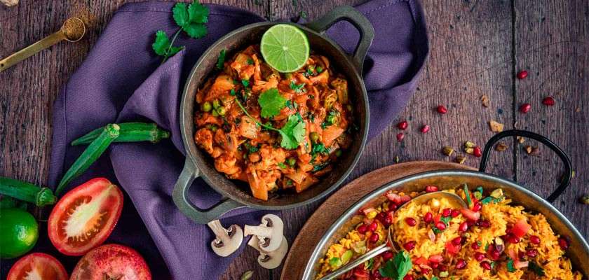 A pot of vegetable curry sits garnished with cilantro and lime, accompanied by a pan of yellow rice. Surrounding are tomatoes, okra, pomegranate, and limes on a wooden tabletop.