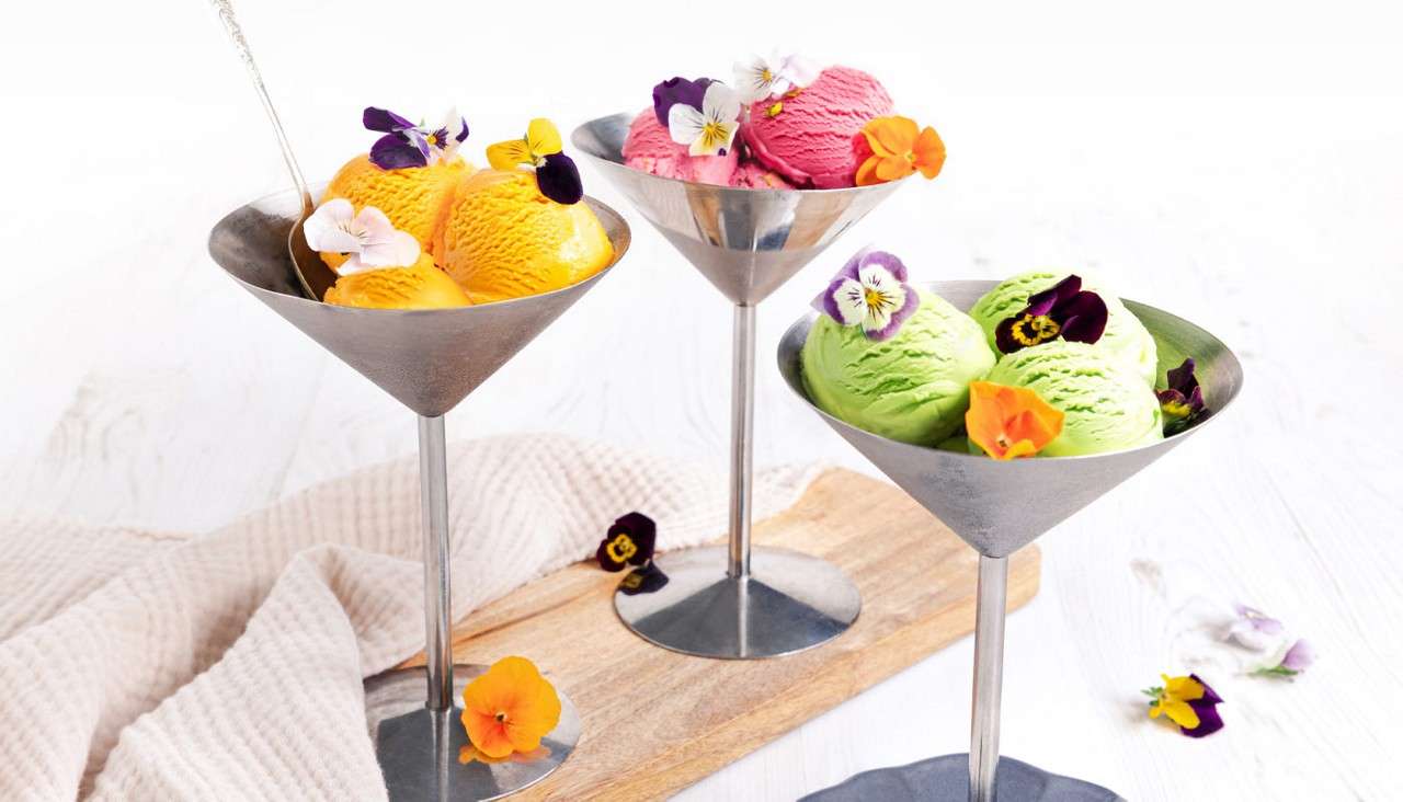 Scoops of colorful ice cream sit garnished with edible flowers in metal martini glasses, arranged on a wooden board amid scattered blooms and cloth on a bright tabletop.